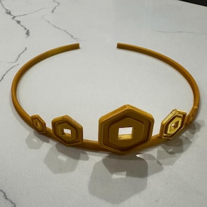 May include: A gold-coloured crown with geometric shapes. The crown features a series of hexagonal and diamond-shaped accents along the front, connected by a thin, curved band. The crown is made of a 3D-printed material.