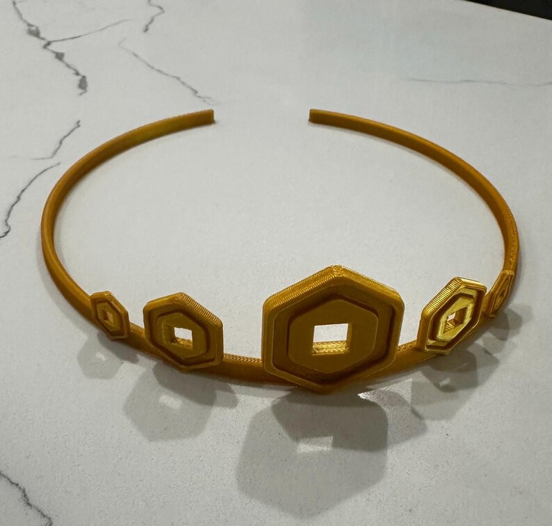 May include: A gold-coloured crown with geometric shapes. The crown features a series of hexagonal and diamond-shaped accents along the front, connected by a thin, curved band. The crown is made of a 3D-printed material.