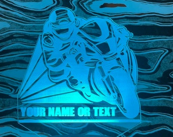 May include: A blue 3D printed night light shaped like a motorcycle racer. The light has a base and a clear acrylic panel with a silhouette of a motorcycle racer. The text "YOUR NAME OR TEXT" is printed on the panel.