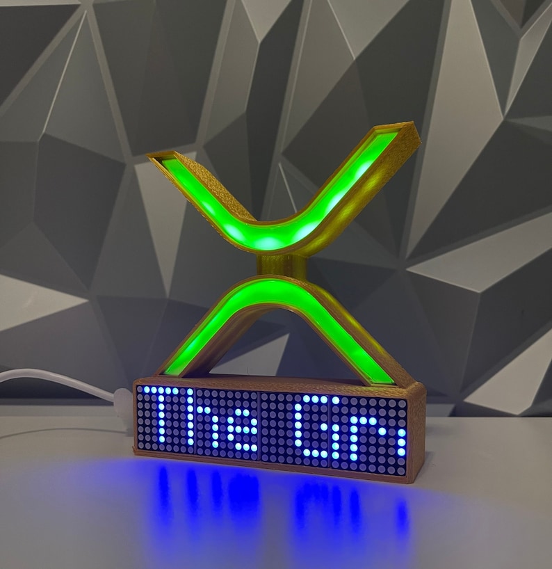 Puede incluir: Una escultura en forma de X, impresa en 3D, de color dorado, con luces LED verdes. La escultura est&aacute; montada sobre una base con una pantalla LED azul que dice "The Lab".