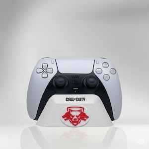 May include: A white and black PlayStation 5 controller resting on a white stand. The stand features the "Call of Duty" logo and a red emblem. The controller has a sleek design with buttons and joysticks.