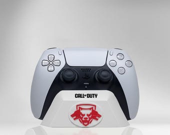 May include: A white and black PlayStation 5 controller resting on a white stand. The stand features the "Call of Duty" logo and a red emblem. The controller has a sleek design with buttons and joysticks.