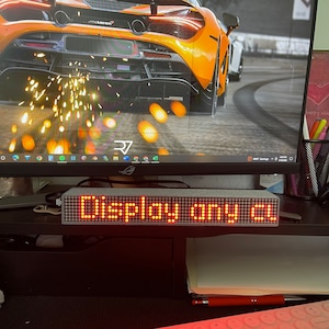 May include: A rectangular LED display with the words "DISPLAY ANY CL" in bright red. The display is on a desk in front of a computer monitor. The monitor displays a car image.