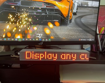 May include: A rectangular LED display with the words "DISPLAY ANY CL" in bright red. The display is on a desk in front of a computer monitor. The monitor displays a car image.