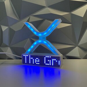 Puede incluir: Un letrero blanco en forma de 'X' impreso en 3D, iluminado con una pantalla LED azul que muestra "The G" sobre una superficie blanca.