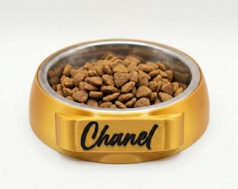 May include: A gold-colored pet food bowl with a stainless steel interior, filled with brown kibble. The name "Chanel" is printed in black on the front of the bowl. The bowl is designed for pets.