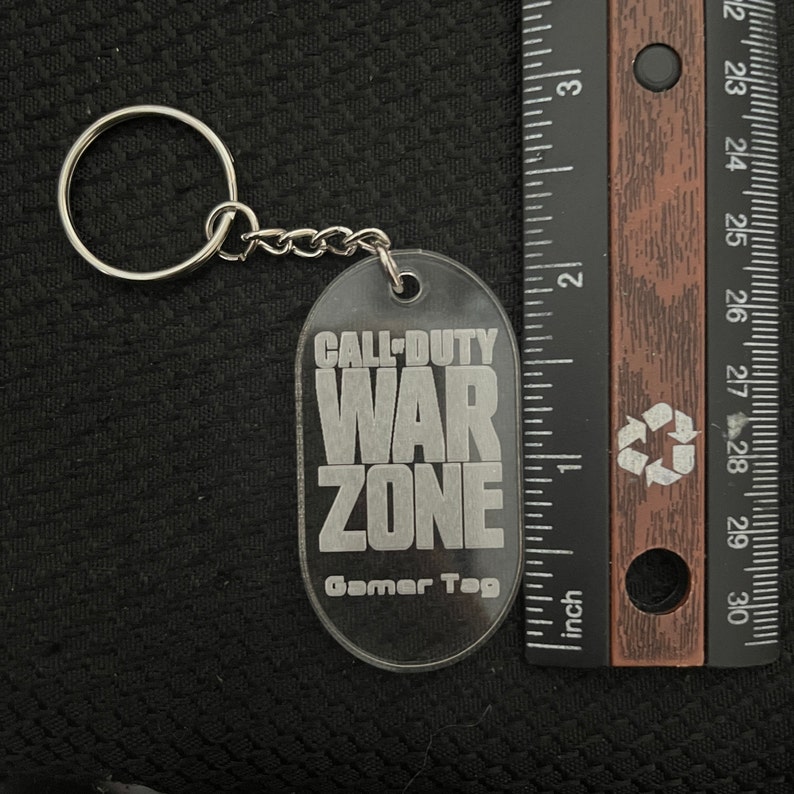 K&ouml;nnte beinhalten: Ein durchsichtiger Schl&uuml;sselanh&auml;nger aus Kunststoff mit einem silbernen Metallring. Der Schl&uuml;sselanh&auml;nger tr&auml;gt die Aufschrift "Call-Duty War Zone Gamer Tag".