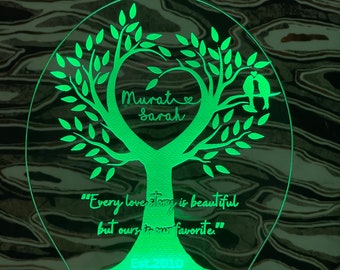 May include: Green illuminated acrylic night light shaped like a tree with a heart in the center. The heart has the names "Murat" and "Sarah" inside. The text below the tree reads "Every love story is beautiful but ours is our favorite." and "Est. 2010".