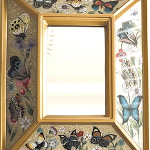 May include: A rectangular gold-colored framed mirror with a decorative border featuring colorful butterflies and flowers. The butterflies have various wing patterns and colors, including yellow, blue, and black. The floral accents include pink, purple, and white blossoms.