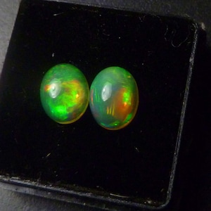 May include: Two oval-shaped opals with a vibrant green base color and internal flashes of orange and yellow. The gemstones are set against a black background, showcasing their iridescent qualities.