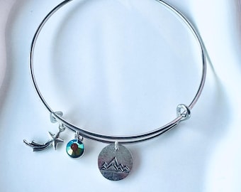 May include: A silver-toned wire bangle bracelet featuring three dangling charms: a shooting star, a small round iridescent crystal bead, and a circular metal disc engraved with a mountain range silhouette design.