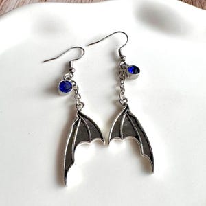 Puede incluir: Un par de pendientes colgantes de tono plateado con dijes de alas de murci&eacute;lago suspendidos de finas cadenas met&aacute;licas. Cada pendiente incluye una peque&ntilde;a piedra redonda de color azul intenso facetada en un engaste plateado cerca del gancho.