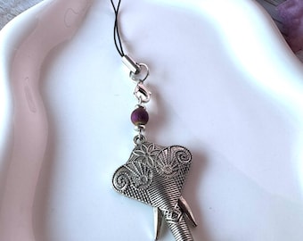 May include: A silver-toned metal elephant charm phone strap featuring intricate etched patterns on the ears and trunk. The charm is attached to a black cord loop with a small, dark purple, textured spherical bead and a silver lobster clasp.