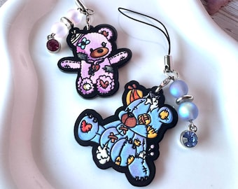 May include: Two phone charms featuring illustrated teddy bears with stitched patches. One bear is pink with a dark red rhinestone, and the other is blue with a light blue rhinestone. Both charms include iridescent beads and black nylon loops.