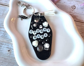 May include: A black coffin-shaped keychain featuring the text SPOOKY BABE in white letter beads. The surface is decorated with small white roses, a skull, a tombstone with RIP, a crystal ball icon, and black rhinestones. It includes a silver ring.