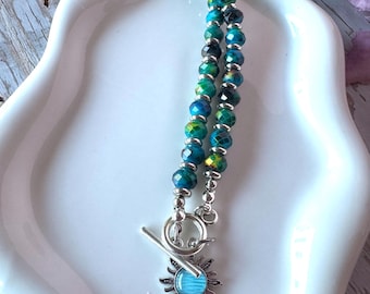 May include: A necklace featuring faceted beads in shades of teal, blue, and yellow, interspersed with small silver-toned metal spacers. The necklace closes with a silver-toned toggle clasp that features a sun-shaped charm with a blue center.