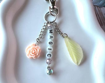 May include: A silver-toned metal keychain featuring a lobster clasp and three dangling charms. One charm is a peach-colored rose, another is a vertical strand of white beads spelling the word MAGIC, and the third is a pale yellow leaf-shaped charm.