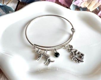 May include: Silver-toned bangle bracelet with three charms. Charms include a leopard, a dragon, and a dark green gemstone. The bracelet is displayed on a white, scalloped dish.