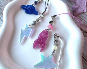 May include: Three phone charms featuring space-themed resin pendants. One charm has a blue glitter planet and a translucent lightning bolt. Another has a pink glitter rocket. The third features a translucent iridescent star with a white cord.