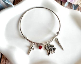 May include: A silver-colored bangle bracelet with multiple charms. Charms include a cross, a red gem, a charm with the words "Bite Me", and a silver spike. The bracelet is displayed on a white, oval-shaped dish.
