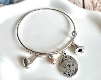 May include: Silver-toned charm bracelet with a pipe, a small pearl, a compass charm, and a horn charm. The compass charm has the text "not all who wander are lost". The bracelet is on a white surface.