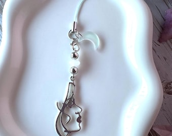 May include: A phone charm featuring a silver-toned metal outline of a human face profile, a small translucent crescent moon charm, and two round beads on a thin cord. The charm is suspended from a clear loop, showcasing a minimalist design.