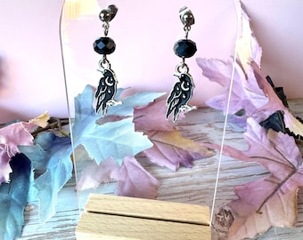 May include: A pair of dangle earrings featuring a black faceted bead and a silver-toned metal charm shaped like a crow with a crescent moon and star detail. The earrings are displayed on a clear acrylic stand with a light wood base.