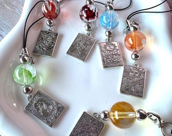 May include: Six phone charms featuring silver-toned rectangular tarot card pendants and colorful glass beads. Each charm includes a black cord loop. The pendants depict various tarot imagery including The Star, The Sun, and The Moon with clear text.