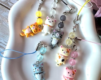 May include: A collection of six cat-shaped charms attached to colorful mobile phone straps. Each charm features a small feline figure in various colors including orange, white, gray, and pink, accented with matching translucent beads and metal hardware.