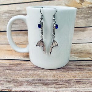 Puede incluir: Un par de pendientes colgantes de tono plateado con una peque&ntilde;a piedra circular azul sobre una cadena de plata que conecta con un dije de ala de murci&eacute;lago. Los pendientes cuelgan de ganchos de metal plateado con un dise&ntilde;o de ala detallado.