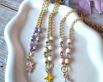 May include: Three gold-tone chain necklaces featuring beads and star-shaped charms. The first necklace has purple beads and a pink star charm, the second has white beads and a yellow star charm, and the third has pink beads and a white star charm.