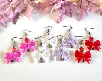 May include: A collection of colorful bow-shaped earrings. The earrings are in shades of pink, white, purple, and red. Each earring has a heart-shaped bead and a small charm. The earrings are displayed on a white surface.