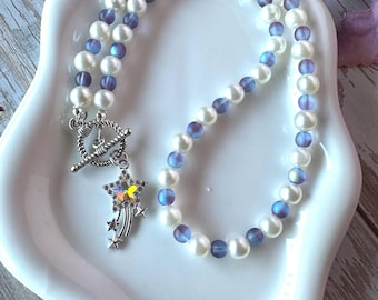 May include: A necklace featuring alternating white faux pearls and translucent purple beads. The piece includes a silver-toned toggle clasp and a dangling charm shaped like a shooting star with a central iridescent stone and smaller star accents.