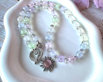 May include: A beaded necklace featuring translucent pastel-colored spheres in shades of pink, blue, and green, interspersed with small clear faceted beads. The necklace includes a silver-toned toggle clasp and a sun-shaped charm with a pink center.