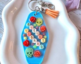 May include: A blue motel-style keychain featuring the text BUENAS VIBRAS spelled out with round letter beads. The surface is decorated with small red rose charms, two skull charms in green and yellow, and tiny colorful beads, with an orange tassel.