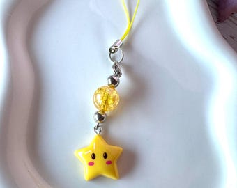 May include: A yellow star-shaped charm featuring a smiling face with pink cheeks, attached to a yellow cord. The charm includes a clear yellow crackle-textured bead and two small silver-toned metallic beads connected by a silver metal jump ring.