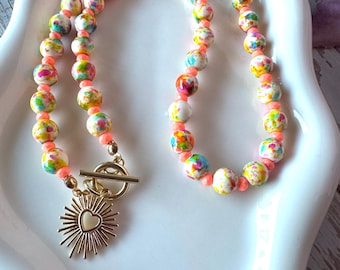 May include: A necklace featuring round beads with a multi-colored speckled pattern in shades of pink, yellow, blue, and green, interspersed with small neon orange spacer beads. It includes a gold-toned toggle clasp and a sunburst heart charm.
