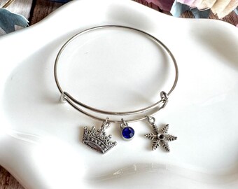 May include: Silver-toned bangle bracelet with three charms: a crown, a blue gem, and a snowflake. The bracelet is displayed on a white, scalloped-edge plate, with a wooden background and decorative leaves.