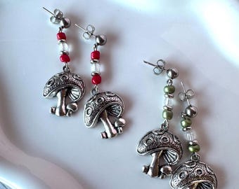 May include: Two pairs of drop earrings featuring silver-toned mushroom charms. One pair includes red and clear beads, while the other pair features olive green and clear beads. Each earring has a metal post back and a dangling mushroom pendant.