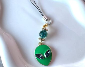 May include: A phone charm featuring a bright green alien head pendant with two dark, oval-shaped crystal eyes. The charm is attached to a black cord and includes a single translucent teal bead accented by two small, metallic silver-toned spacer beads.