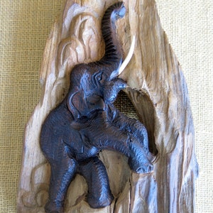 K&ouml;nnte beinhalten: Eine dunkle Holzschnitzerei eines Elefanten mit erhobenem R&uuml;ssel und einem Sto&szlig;zahn. Der Elefant ist realistisch geschnitzt und vor einem Hintergrund aus geschnitztem Holz platziert.