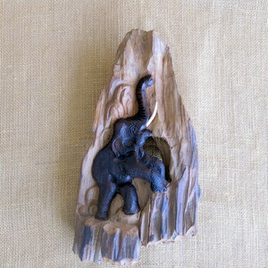 K&ouml;nnte beinhalten: Eine geschnitzte Elefantenfigur aus Holz mit erhobenem R&uuml;ssel und Sto&szlig;z&auml;hnen. Der Elefant ist dunkelbraun und vor einem hellbraunen Holzhintergrund platziert.