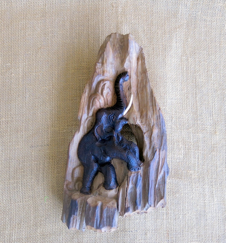 K&ouml;nnte beinhalten: Eine geschnitzte Elefantenfigur aus Holz mit erhobenem R&uuml;ssel und Sto&szlig;z&auml;hnen. Der Elefant ist dunkelbraun und vor einem hellbraunen Holzhintergrund platziert.