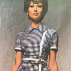 Pode incluir: Uma mulher vestindo um vestido de manga curta com um padrão xadrez branco e preto. O vestido tem uma bainha branca e um cinto branco com fivela. O vestido tem uma faixa branca que atravessa diagonalmente a frente do vestido.