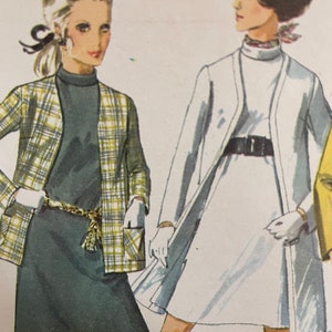 May include: Two women wearing vintage 1960s style dresses. The woman on the left is wearing a plaid jacket over a black dress with a belt. The woman on the right is wearing a white dress with a black belt and a white jacket.