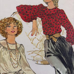 May include: Two women wearing blouses with long sleeves and a gathered neckline. The woman on the left is wearing a white blouse with a beaded necklace and a brown belt. The woman on the right is wearing a red blouse with a black and gold print and a brown belt.