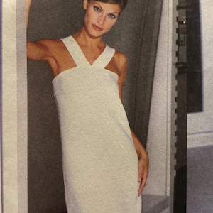 May include: A white dress with a distinctive neckline and shoulder straps. The dress has a simple, elegant design and appears to be made of a smooth fabric. The model is wearing the dress in a studio setting.