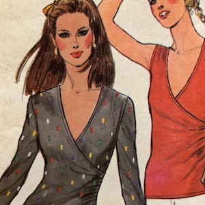 Op de afbeelding: Een mode-illustratie met twee tops. De ene is een donkergrijze wikkel-top met lange mouwen en een patroon van kleine, veelkleurige rechthoekige streepjes. De andere is een mouwloze, effen rode wikkel-top met een diepe V-hals.