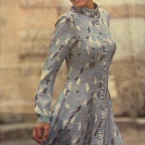 May include: A woman wearing a long-sleeved dress with a blue and white abstract pattern. The dress has a high collar and a button-up front.