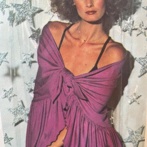 May include: A woman with curly brown hair is wearing a purple, ruffled top with a black bra strap showing. She is standing in front of a silver sparkly background with stars.